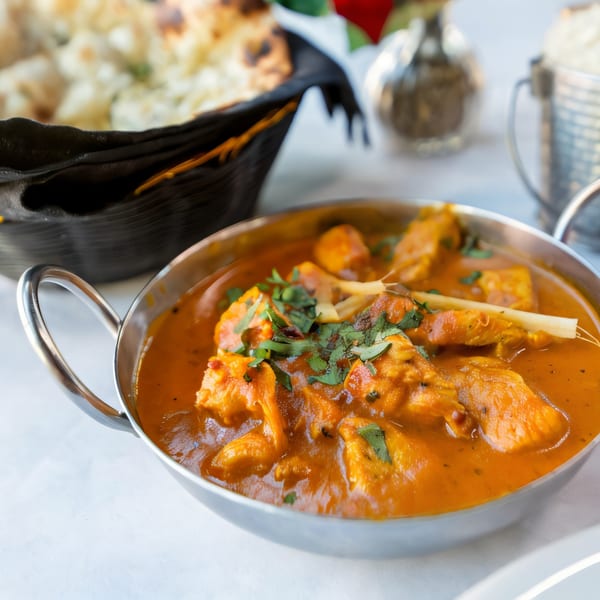 Indian Oven's chicken curry in a metal bowl, garnished with cilantro, with naan bread in the background.