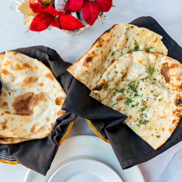 Two baskets of Indian naan bread, one plain and one with herbs and seeds, on a white table.