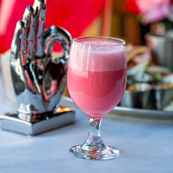 A vibrant pink Indian lassi drink in a stemmed glass, with a silver hand sculpture in the background.